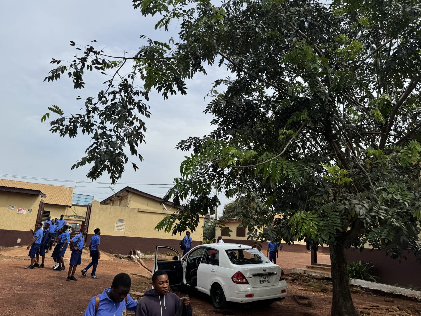 Students in blue uniform arriving on a school compound under trees