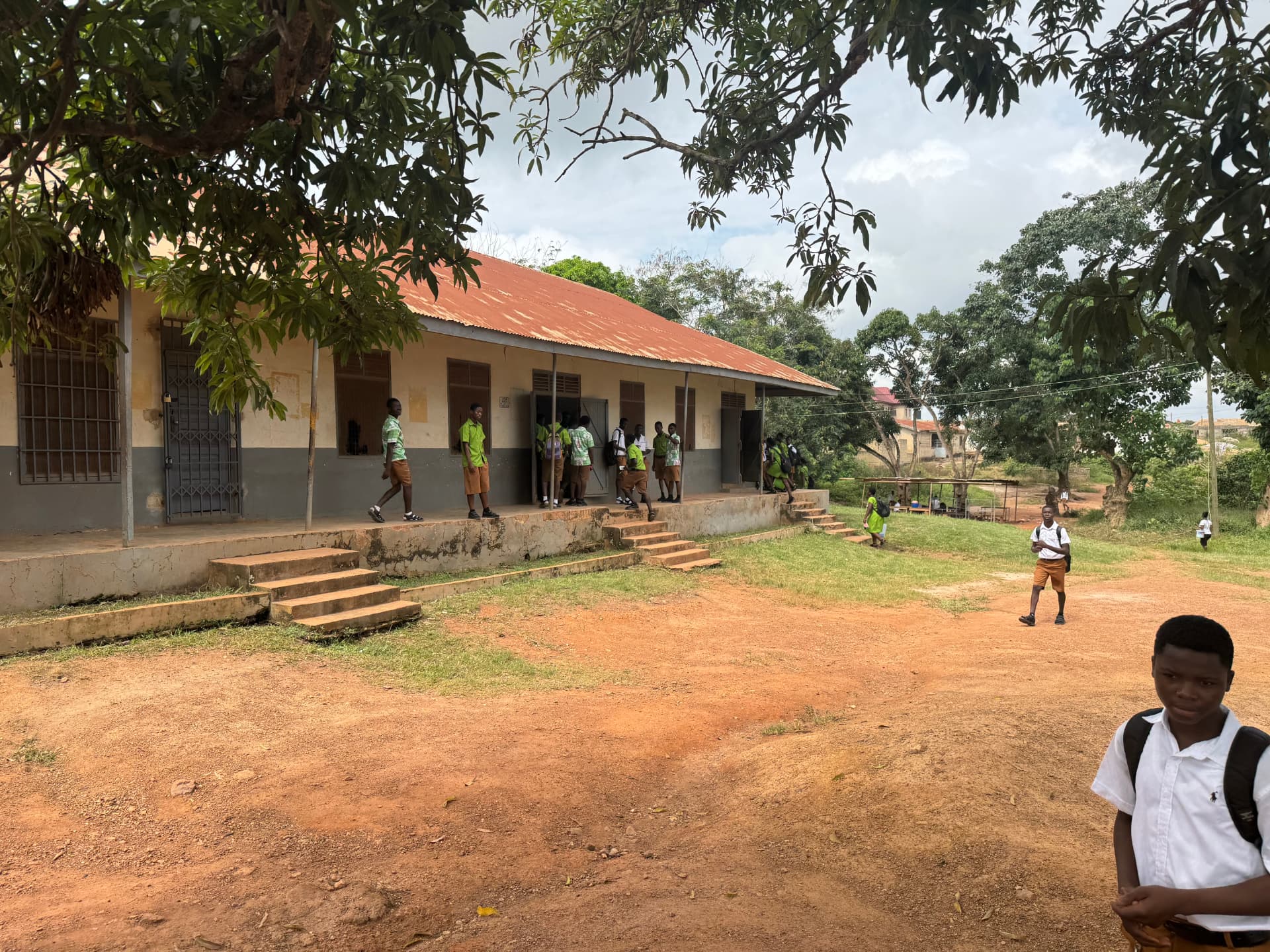 Students in green and brown uniforms outside a basic school block with a red roof