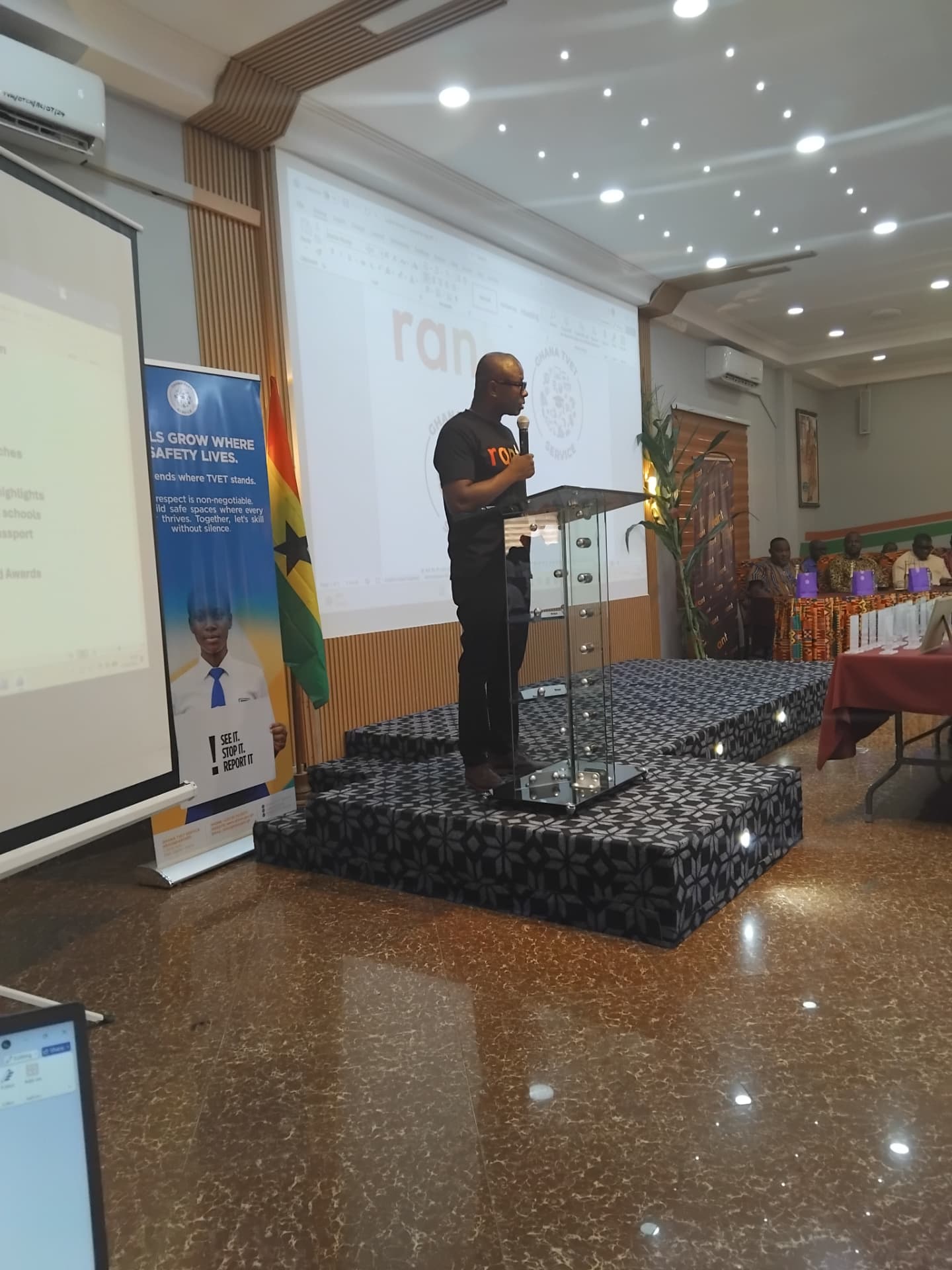 Speaker at a podium on stage with the RANT logo projected and a Ghana flag beside a safeguarding banner at the Tru Vine Hotel, Kumasi