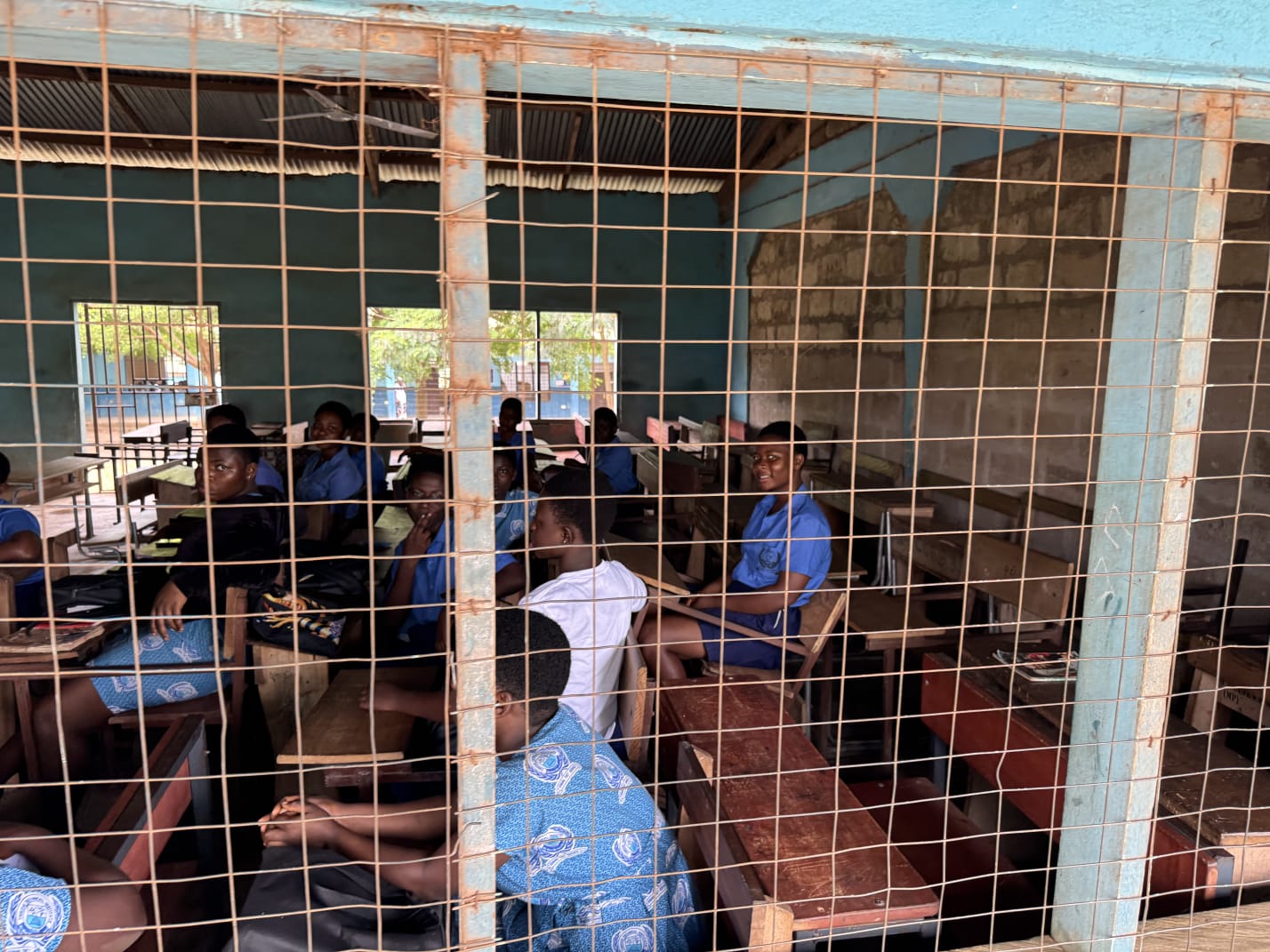 Students in blue uniform seated at wooden desks in an open-sided classroom in Ghana